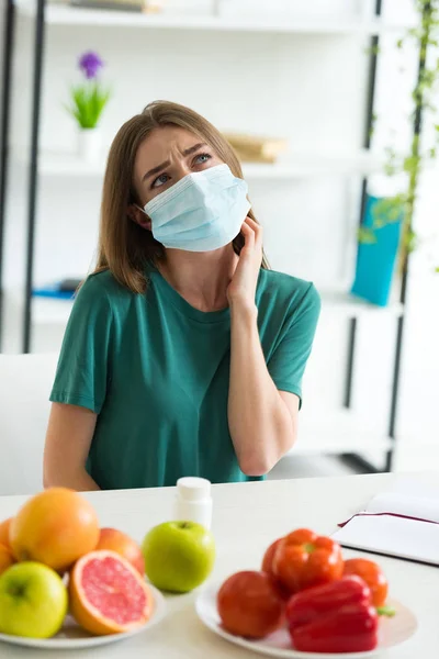 Задумчивая женщина в медицинской маске сидит за столом с фруктами, овощами и таблетками дома — стоковое фото