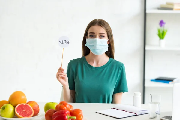 Вид женщины в медицинской маске, сидящей за столом с фруктами, овощами и таблетками и держащей баннер на палочке с надписью аллергия на дома — стоковое фото