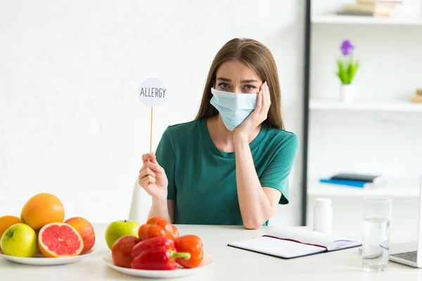 Mujer en máscara médica sentada a la mesa con frutas, verduras y pastillas y sosteniendo pancarta en palo con alergia a la inscripción en el hogar - foto de stock