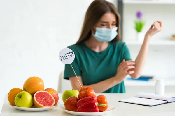 Foco seletivo da mulher na máscara médica coçar enquanto se senta à mesa com legumes, frutas, pílulas e modelo com alergia à inscrição — Fotografia de Stock