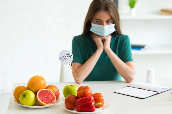 Грустная женщина в медицинской маске подпирает лицо руками, сидя за столом с фруктами, овощами и шаблоном с надписью аллергия — стоковое фото