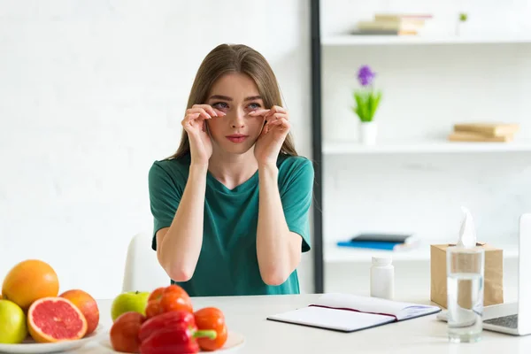Девушка с аллергией вытирать слезы, сидя за столом с фруктами, овощами и таблетками — стоковое фото