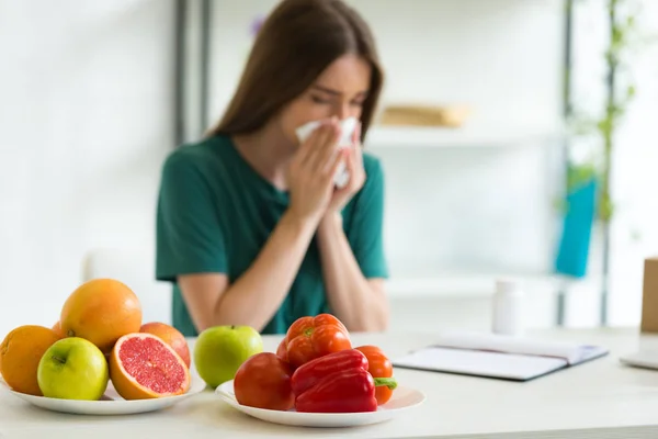 Selective focus of woman sitting at table with vegetables, fruits and pills and using napking while blowing nose — Stock Photo