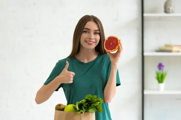 Lächelnde junge Frau hält geschnittene Grapefruit neben Papiertüte mit Lebensmitteln und zeigt Daumen hoch zu Hause — Stockfoto