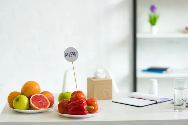 Fresh fruits, vegetables, glass of water, pills, textbook and template with inscription allergy on table — Stock Photo