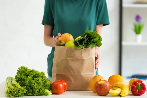 Обрезанный вид женщины со свежими овощами и фруктами в бумажном пакете на столе — стоковое фото