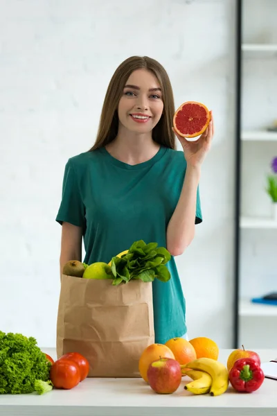 Sorrindo jovem mulher de pé perto saco de papel com legumes frescos e frutas e segurando toranja cortada em casa — Fotografia de Stock