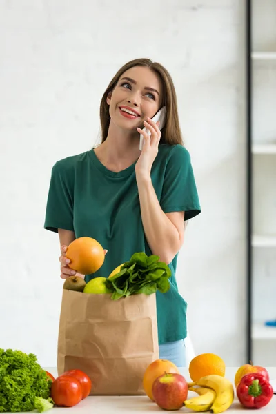 Lächelndes Mädchen, das neben einer Papiertüte mit frischen Lebensmitteln steht, Orange in der Hand hält und zu Hause mit dem Smartphone spricht — Stockfoto