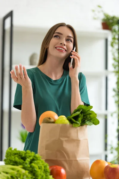 Smiling girl standing near paper bag with fresh food and talking on smartphone at home — Stock Photo