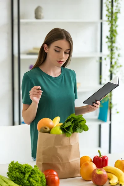 Atractiva mujer sosteniendo pluma y libro de texto mientras está de pie cerca de la bolsa de papel con frutas y verduras frescas - foto de stock