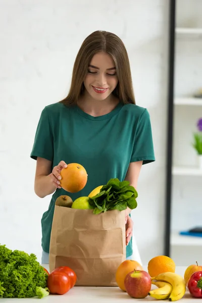 Sonriente joven mujer de pie cerca de bolsa de papel con verduras frescas y frutas en casa - foto de stock