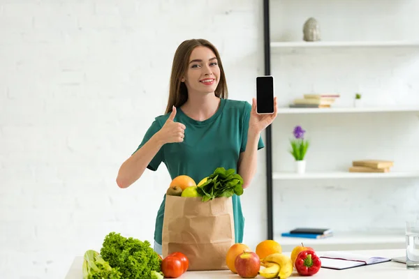 Chica sonriente de pie cerca de la bolsa de papel con comida y la celebración de teléfono inteligente con pantalla en blanco en casa - foto de stock