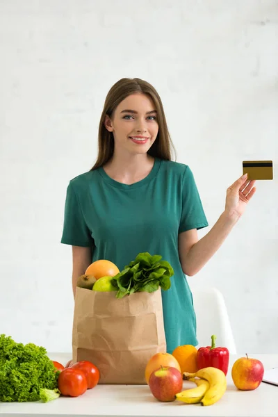 Chica sonriente de pie cerca de la bolsa de papel con comida fresca y la celebración de la tarjeta de crédito en casa - foto de stock