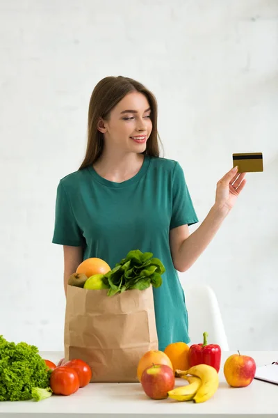 Lächelndes Mädchen steht neben Papiertüte mit frischen Lebensmitteln und hält Kreditkarte zu Hause — Stockfoto