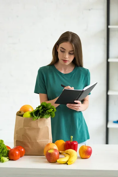 Atractiva mujer sosteniendo pluma y libro de texto mientras está de pie cerca de la bolsa de papel con frutas y verduras frescas - foto de stock