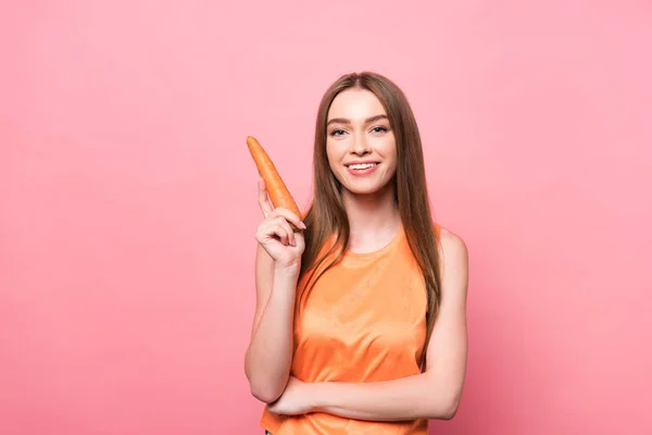 Усміхнена приваблива молода жінка тримає моркву і дивиться на камеру на рожевому — стокове фото