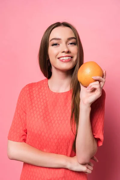 Attraktive lächelnde junge Frau mit Grapefruit und Blick in die Kamera auf rosa — Stockfoto