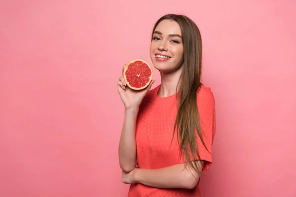 Atractiva joven sonriente sosteniendo toronja cortada y mirando a la cámara en rosa - foto de stock