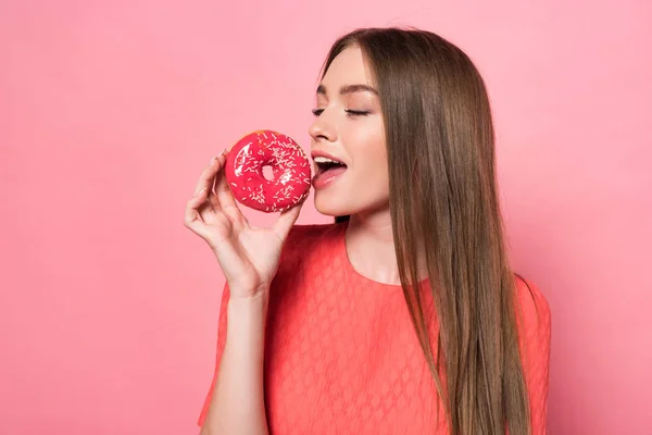 Усміхнена приваблива молода жінка їсть солодкий пончик на рожевому — стокове фото