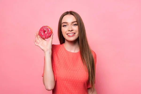 Вид спереду усміхненої привабливої молодої жінки, що тримає солодкий пончик і дивиться на камеру на рожевому — стокове фото