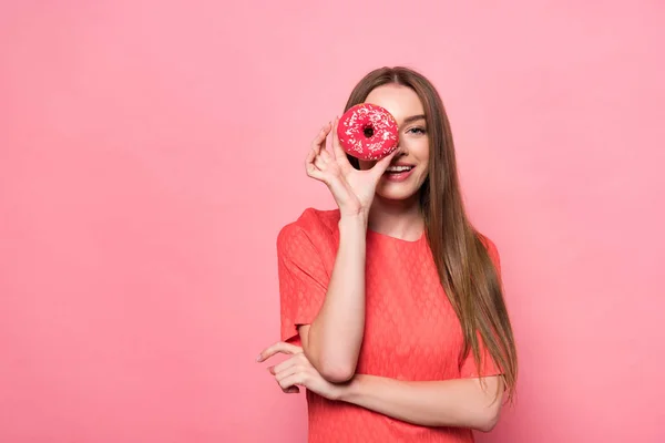 Vue de face de sourire attrayant jeune femme tenant beignet doux et regardant la caméra sur rose — Photo de stock
