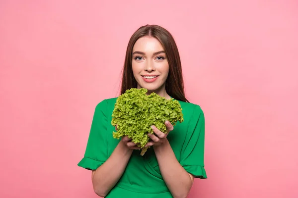Vorderansicht der attraktiven lächelnden jungen Frau mit Salat und Blick in die Kamera isoliert auf rosa — Stockfoto