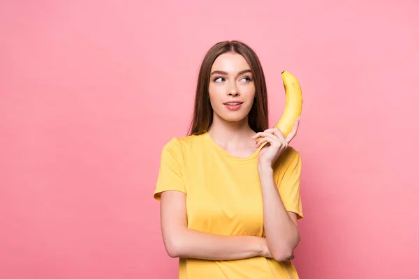 Attractive smiling girl holding banana and looking away on pink — Stock Photo