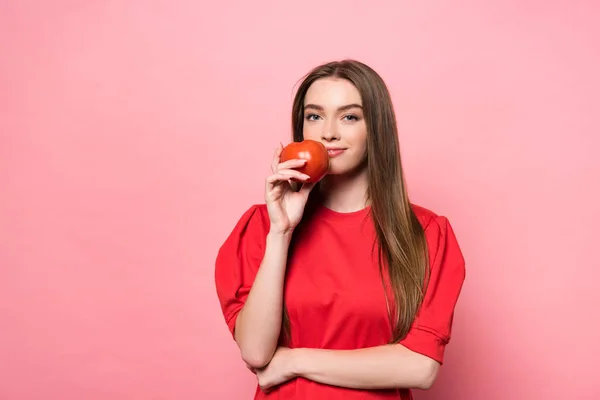 Atractiva chica sonriente sosteniendo tomate y mirando a la cámara en rosa - foto de stock