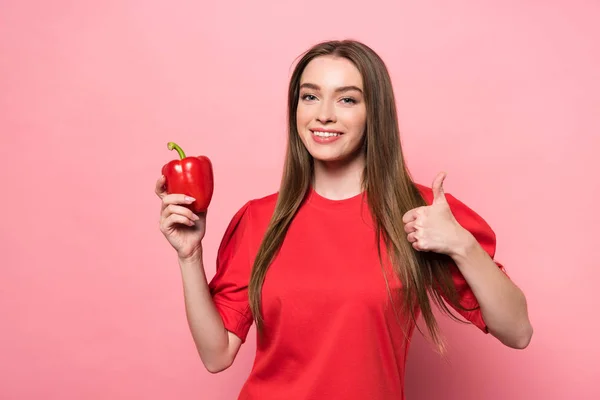 Sonriente joven atractiva sosteniendo pimiento rojo y mostrando el pulgar hacia arriba en rosa - foto de stock