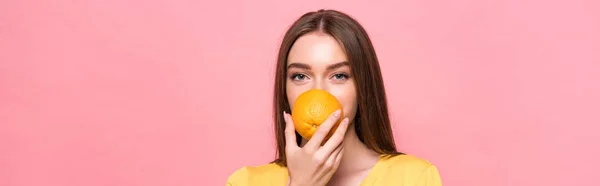 Plano panorámico de la joven mujer comiendo naranja y mirando a la cámara aislada en rosa - foto de stock