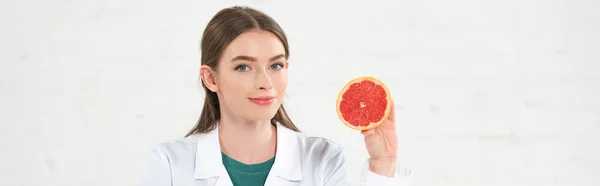Panoramic shot of smiling dietitian in white coat holding cut grapefruit and looking at camera isolated on white — Stock Photo
