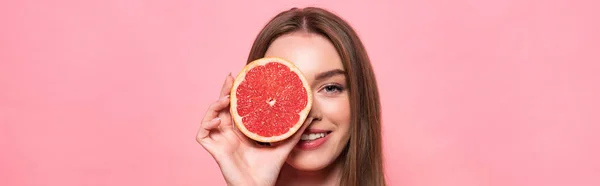 Панорамний знімок усміхненої привабливої дівчини, що тримає розрізаний грейпфрут ізольований на рожевому — стокове фото