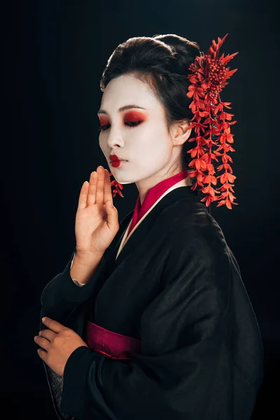 Bela gueixa em quimono preto e vermelho e flores no cabelo com a mão perto do rosto isolado em preto — Fotografia de Stock