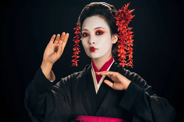 Schöne Geisha in schwarzem und rotem Kimono und Blumen im Haar, die isoliert auf schwarz gestikulieren — Stockfoto