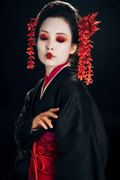 Hermosa geisha en kimono negro y rojo y flores en el pelo mirando hacia abajo aislado en negro - foto de stock