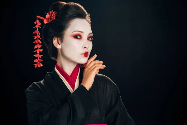 Hermosa geisha en kimono negro y rojo y flores en el pelo con la mano cerca de la cara aislada en negro - foto de stock