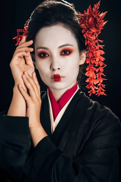 Bela gueixa em quimono preto e vermelho e flores no cabelo com as mãos perto do rosto isolado em preto — Fotografia de Stock