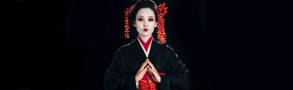 Beautiful geisha in black and red kimono and flowers in hair with clenched hands isolated on black, panoramic shot — Stock Photo