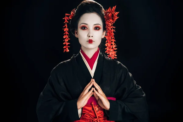 Hermosa geisha en kimono negro y rojo y flores en el pelo con las manos apretadas aisladas en negro - foto de stock
