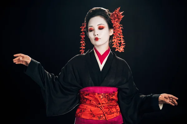 Beautiful geisha in black and red kimono and flowers in hair looking down and gesturing isolated on black — Stock Photo