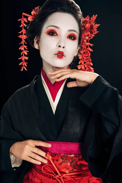 Hermosa geisha en kimono negro y rojo y flores en el pelo tocando la barbilla aislado en negro - foto de stock