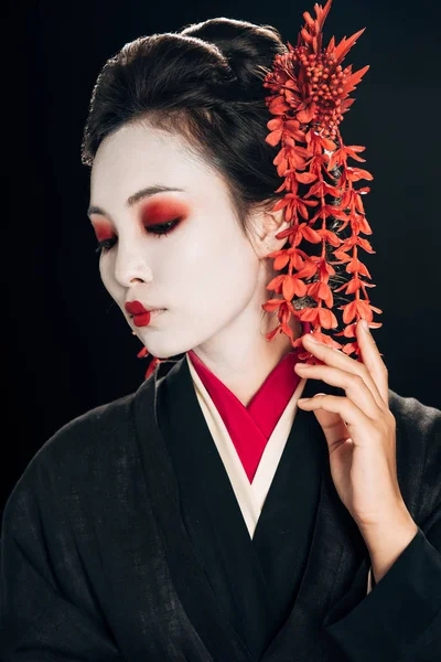 Retrato de bela gueixa em quimono preto e vermelho e flores no cabelo olhando para baixo isolado em preto — Fotografia de Stock