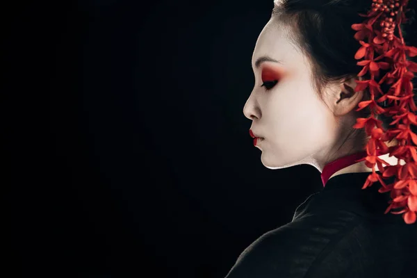 Perfil de hermosa geisha en kimono negro y rojo y flores en pelo aislado en negro con espacio de copia - foto de stock