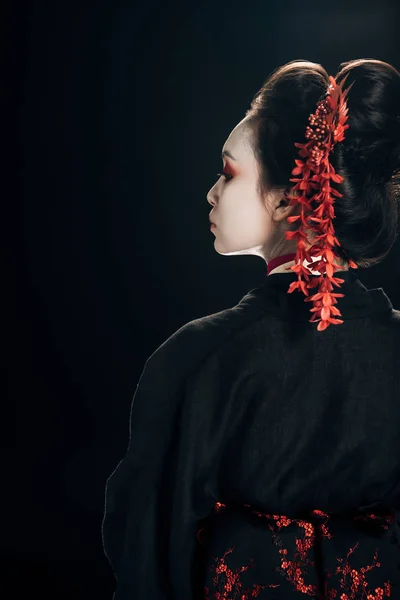 Back view of beautiful geisha in black and red kimono and flowers in hair looking away isolated on black — Stock Photo