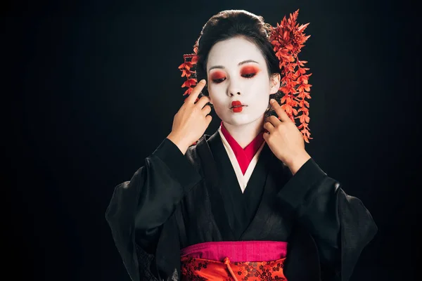 Hermosa geisha en kimono negro y rojo y flores en el pelo tocando la cara aislado en negro - foto de stock