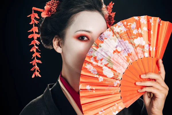Hermosa geisha con flores rojas en el pelo sosteniendo ventilador de mano tradicional aislado en negro - foto de stock