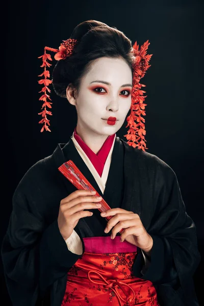 Belle geisha en kimono noir avec des fleurs rouges dans les cheveux tenant ventilateur main traditionnelle isolé sur noir — Photo de stock