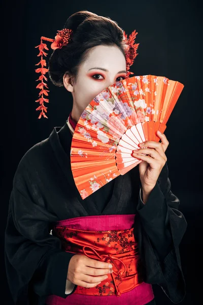 Geisha en kimono negro con flores rojas en el pelo ocultando la cara detrás de ventilador de mano tradicional aislado en negro - foto de stock