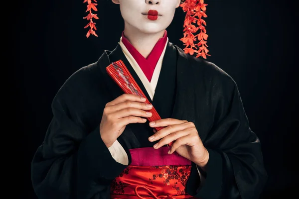 Partial view of geisha in black kimono with red flowers in hair holding traditional hand fan isolated on black — Stock Photo