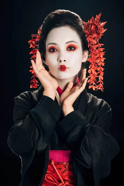 Geisha en kimono noir et rouge et fleurs dans les cheveux avec la main près du visage isolé sur noir — Photo de stock